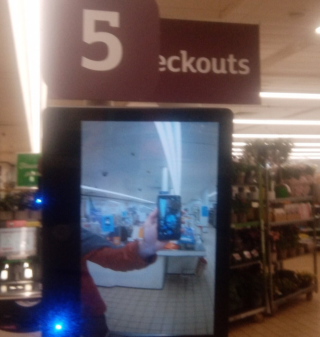 A screen mounted above a supermarket self-checkout lane, showing live footage of the shopper standing in front of it