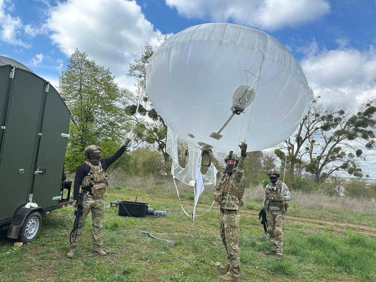 Three Ukrainian soldiers in camouflage stand on grassland holding the tether lines of a white helium aerostat as it rises. A sensor and antenna payload hangs on a strut beneath the envelope, and a green ground trailer carrying the winch sits at the left of the frame