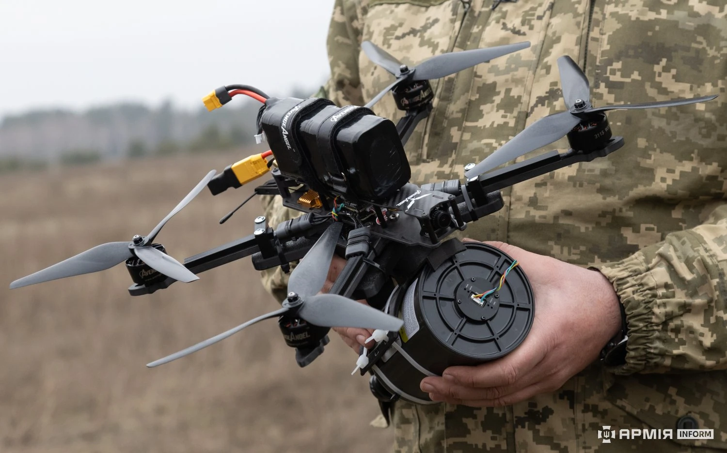 A Ukrainian soldier holds a fibre-optic FPV drone in one hand. The black cylindrical spool canister slung under the airframe is about as large as the drone's own body, and the fibre pays out through a port in its base